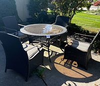 Full view of round wicker patio dining table with glass top and 4 matching wicker chairs with striped cushions placed outdoors on a concrete surface.