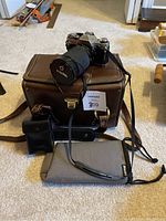 Canon AE-1 camera with attached lens sitting on brown leather carrying case. Also showing Canon Speedlite flash, camera strap, two gray padded bags from Black's.