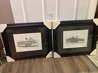 Two black framed prints of Boblo boats, both with protective corner covers visible, placed side by side on wood floor.