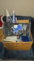 Photo showing a box filled with Detroit Lions books, autograph book, NFL magazines. Also includes a black chair cover with Detroit Lions logo and two white and blue Toronto Blue Jays pennants.