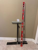 Adjustable roller stand and red spirit level tool shown leaning against wall, both tools placed side by side.