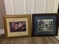 Two framed paintings side by side on floor; left is floral with gold frame, right is farmhouse scene with dark brown frame