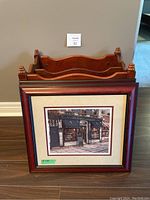 Wooden Bombay-style magazine rack positioned behind two framed wall art pieces on a wooden floor against a beige wall.
