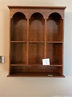 Front view of wooden wall shelf showing three vertical sections and six smaller shelves underneath, decorative arches at top of each vertical section, medium brown wood finish.