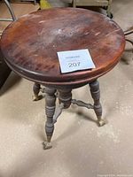 View of the wooden stool from a slightly elevated angle showing the round seat and three of four turned legs ending in metal claw feet.