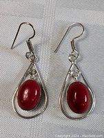 Pair of sterling silver dangle earrings with red oval cabochon stones set in teardrop frames on white fabric background.