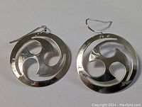 Pair of sterling silver circular earrings with hook wires shown against a plain background; abstract swirl cut-out design visible.