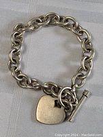 Full view of the sterling silver thick chain bracelet with heart charm and toggle clasp arranged in a circle on a textured fabric surface.