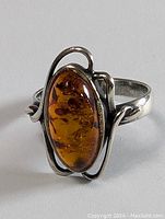 Front and side view of a sterling silver ring featuring a polished oval amber stone with natural inclusions and warm amber tones set in a stylized silver mounting with curved bands.