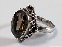 Side angle close-up of a sterling silver ring with an oval faceted orange-tone iolite gemstone surrounded by a decorative cluster bead setting. Marked 925 on the band.