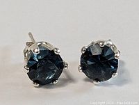 Close-up front angle of pair of sterling silver stud earrings with dark blue stones in six-prong settings.