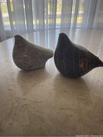 Two stone bird sculptures, one black with light streaks from Italy and one grey speckled from Vancouver, on marble table in front of window.