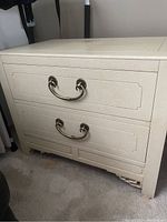 Front side of one off white wooden end table showing two drawers with gold handles and decorative recessed paneling.