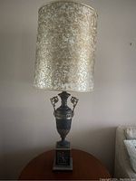 Full view of one table lamp showing black base with gold accents, ornate handles, and cylindrical mixed gold textured shade.