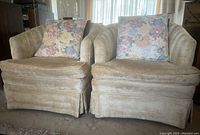 Pair of beige upholstered armchairs with floral cushions on each, showing slight wear.