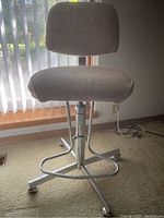 Front-side view of an adjustable art table chair showing gray cushioned seat and backrest, metal base with five legs on caster wheels and circular footrest.