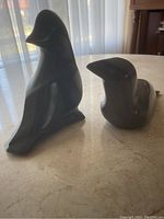 Front and side view of two dark grey seal sculptures, one larger and one smaller, placed on a light-colored surface near a window with sheer curtains.