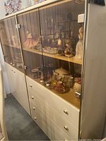 Photo showing cream china cabinet with glass upper doors, shelves with items inside, drawers below, and metal knobs