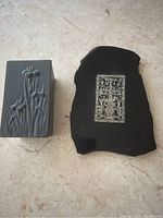 Photo of two decorative items on a light surface: left is a rectangular metal container with iris engravings, right is a black marble piece with a central engraved metal plate.