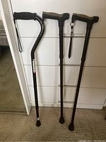 Three black adjustable walking canes, two with T-shaped handles and one with a curved handle, all with rubber grips and wrist straps standing against a white dresser.