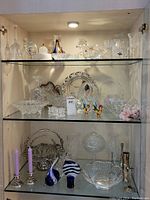 Glass cabinet with three shelves displaying assorted crystal and glass bowls, decanters, glasses, and figurines.