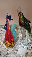 Both glass bird figurines placed side by side on a countertop, showing the rooster and cockatoo fully.