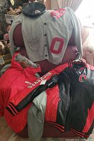 Photo showing all items together on a chair: grey cardigan with red O, black leather cap, red and grey jacket, and black/red athletic jacket