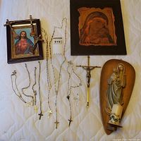 Full lot showing framed Sacred Heart of Jesus photo, Madonna and Child icon, wood carving with votive holder, rosaries and crucifixes laid out on white quilt.