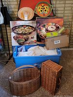 Group photo showing the boxed Fiesta ceramic serving sets, cowboy hat chip and dip bowl, Italian deep dish, blue tote with linens, and two woven baskets on floor against brick wall.