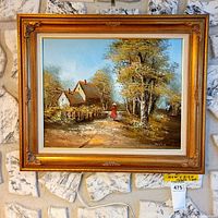 Framed oil painting hanging on a stone wall, depicting a woman walking down a road beside cottages and trees, with gilded frame and linen matting.