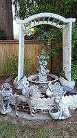 White plastic pergola arch standing over central fountain basin with assorted statues at its base.
