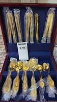 Goldtone flatware set arranged in maroon storage box with blue lining. Pieces individually wrapped in plastic for protection.