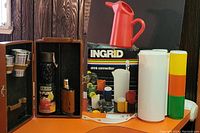 Photo showing wooden travel bar case containing bottle, flask, metal cups, and shot glasses, with Ingrid Drink Connection box, stacked colorful cups, and white pitcher beside it.