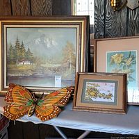 Oil painting of mountain scene with river and cabin, large wooden butterfly, framed 3D floral and stagecoach pictures on table