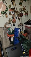 Wall with hanging various packaged small hardware items and tools, green golf bag, blue camping bag, and hand tools on floor