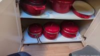 Wide view of red enamel cookware including cast iron pans with lids, roaster, stock pots, and oval casserole dish on cabinet shelves.