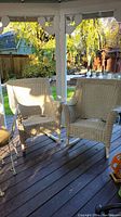 Two cream-colored wicker rocking chairs arranged side by side on outdoor porch with wood flooring and garden background.