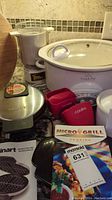 Photo showing Rival Crock Pot slow cooker with glass lid, Cuisinart waffle maker, Rival Little Dipper, Micro Grill boxed, two red silicone grabbers, and wooden round cutting board.