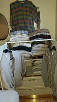 Photo of stacked sweaters on white dresser, showing assortment of folded sweaters and one hanging multicolor checked crewneck sweater