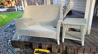 White wicker settee and side table on outdoor deck with grass in background. Setee has curved back and armrests, side table has square top and lower shelf.