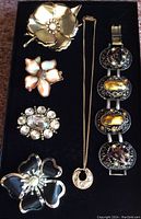 Full view of all jewelry items arranged on black background showing the four brooches in various floral styles, gold-tone necklace with circular pendant, and decorative bracelet with large amber stones and rhinestones.