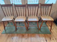 Front view of four matching antique wooden chairs with spindle backs and contoured seats, arranged side by side on a green rug.