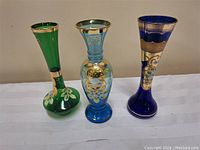 Three Bohemian hand blown art glass vases in green, light blue, and cobalt blue with gold trim and floral enamel decoration shown together.