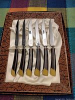 Six vintage style serrated cutting knives with wood-look handles and beige tips, arranged inside satin-lined presentation box with textured brown exterior.