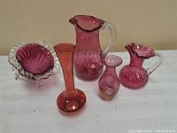 Front view of five cranberry glass items including pitchers, vases, and bowl arranged on white surface