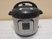 Front view of the Instant Pot showing its stainless steel body, black lid with handle, and digital control panel.