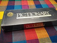 Front top side of the Pictionary game box showing the brand name, title, and first edition label on a colorful background.