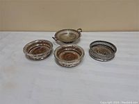 Four items: three silver plated round wine bottle coasters of similar design and one round silver plated serving tray with two small handles, laid on a white surface.
