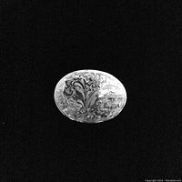 Close-up black and white photo of an oval brooch showing detailed floral embossing on the silver surface against black background.