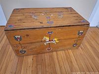 Front view of wooden toy storage trunk with hand-painted folk art including a carousel horse and floral patterns on a natural wood finish surface, showing wood grain and signs of use.